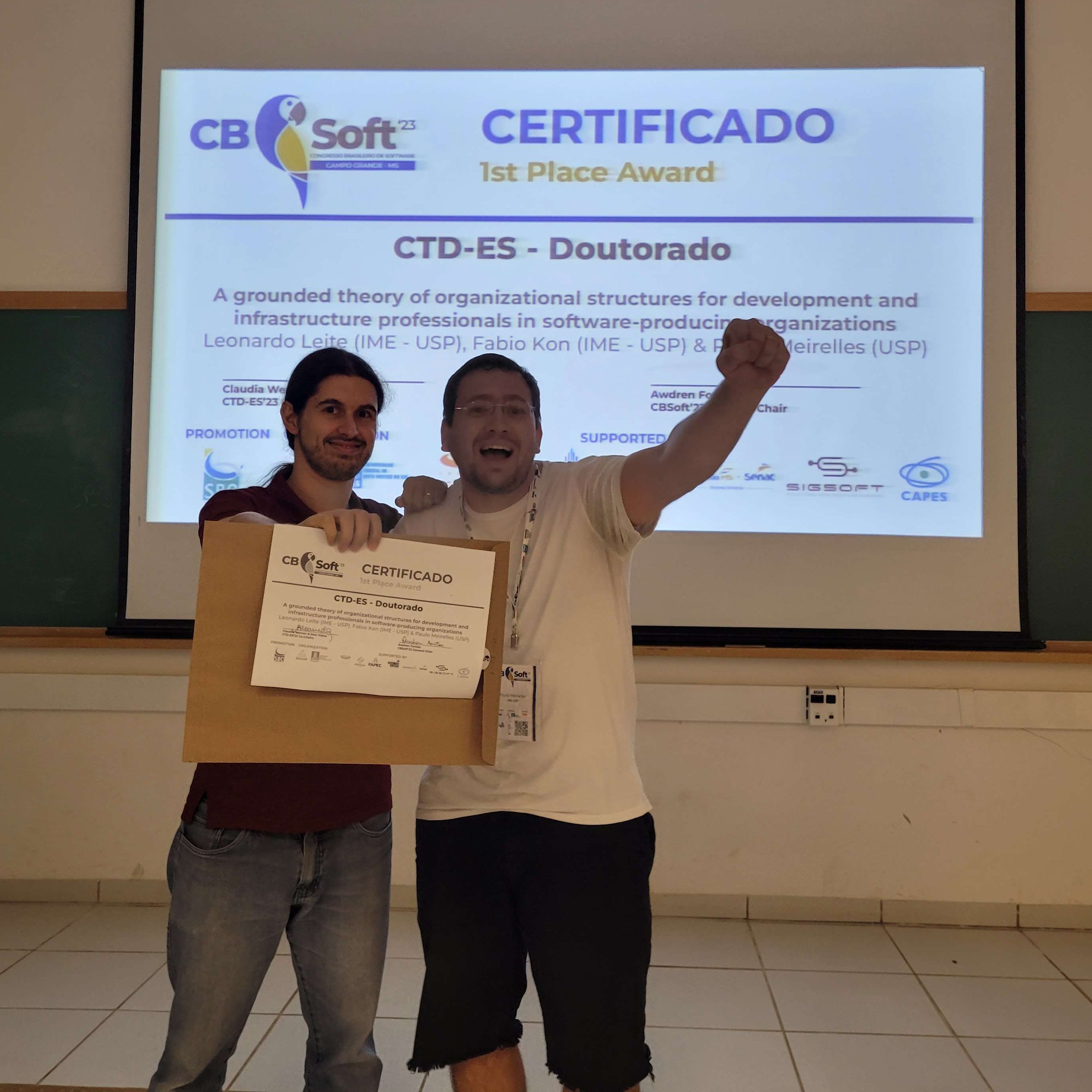 Leonardo e Paulo segurando o certificado; Paulo comemorando com punho erguido; atrás telão exibindo certificado da premiação.